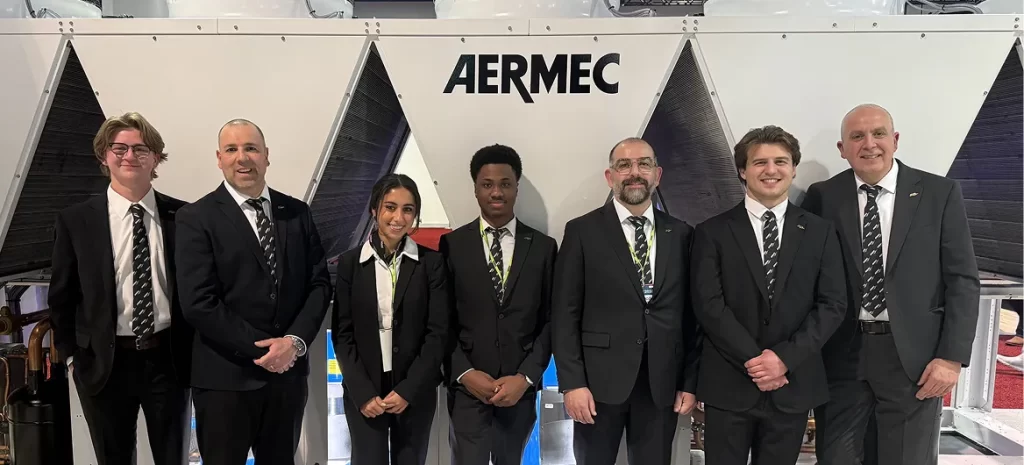 Group of seven professionals in black suits posing in front of an AERMEC display at a trade show or facility entrance.