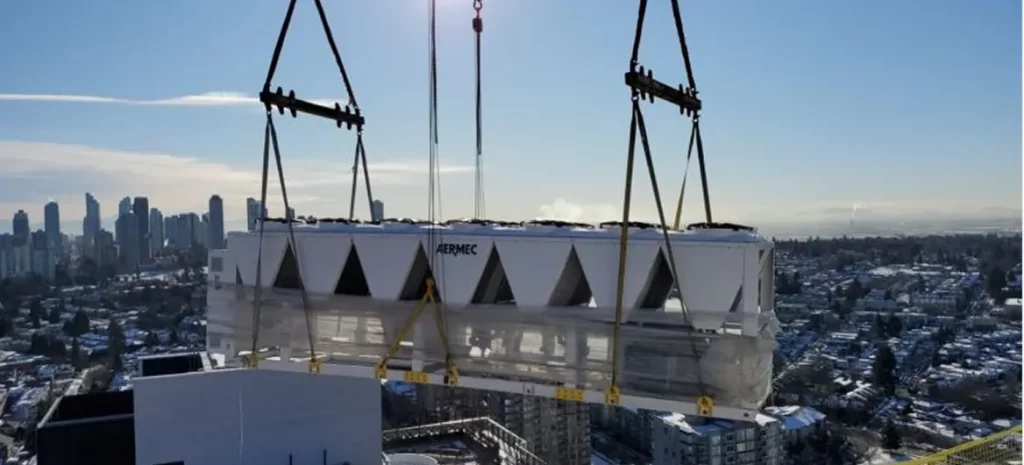 Industrial HVAC unit being hoisted onto a rooftop with cranes, city skyline in the distance.