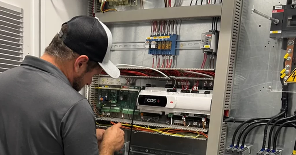 Electrical technician wiring inside an industrial control panel, with a COS+ device mounted and various wires exposed