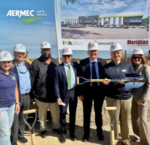Eight people wearing hard hats pose together at a construction site, holding a ceremonial shovel with a branded banner in the background.