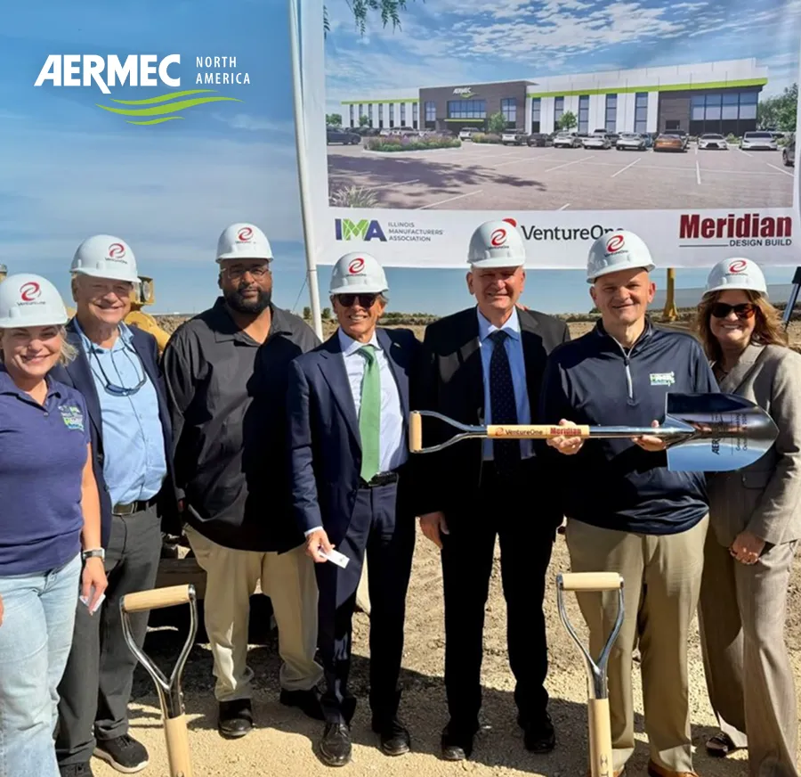 Eight people wearing hard hats pose together at a construction site, holding a ceremonial shovel with a branded banner in the background.